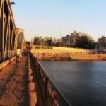 Pasarela de un puente sobre el Nilo en El Cairo al atardecer, con peatones, palmeras y edificios modernos de la ciudad iluminados por luz calida