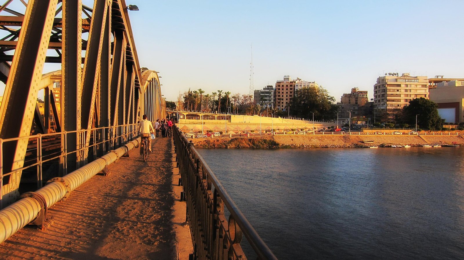 Pasarela de un puente sobre el Nilo en El Cairo al atardecer, con peatones, palmeras y edificios modernos de la ciudad iluminados por luz calida