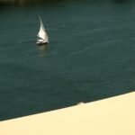 Felucca de vela blanca navegando por las aguas turquesa del rio Nilo junto a un banco de arena dorado en Egipto, escena ideal para preparar un viaje saludable