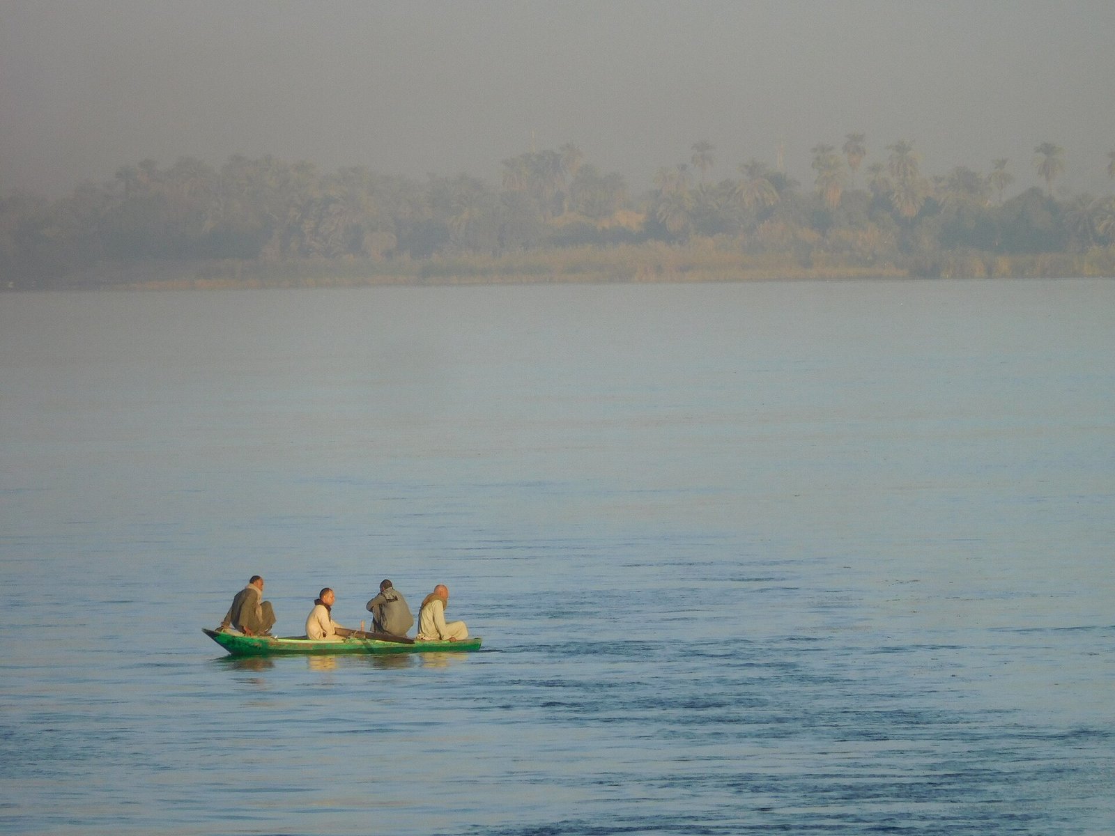 Pequena barca de madera con pescadores locales sobre el rio Nilo al amanecer brumoso cerca de palmeras en Egipto