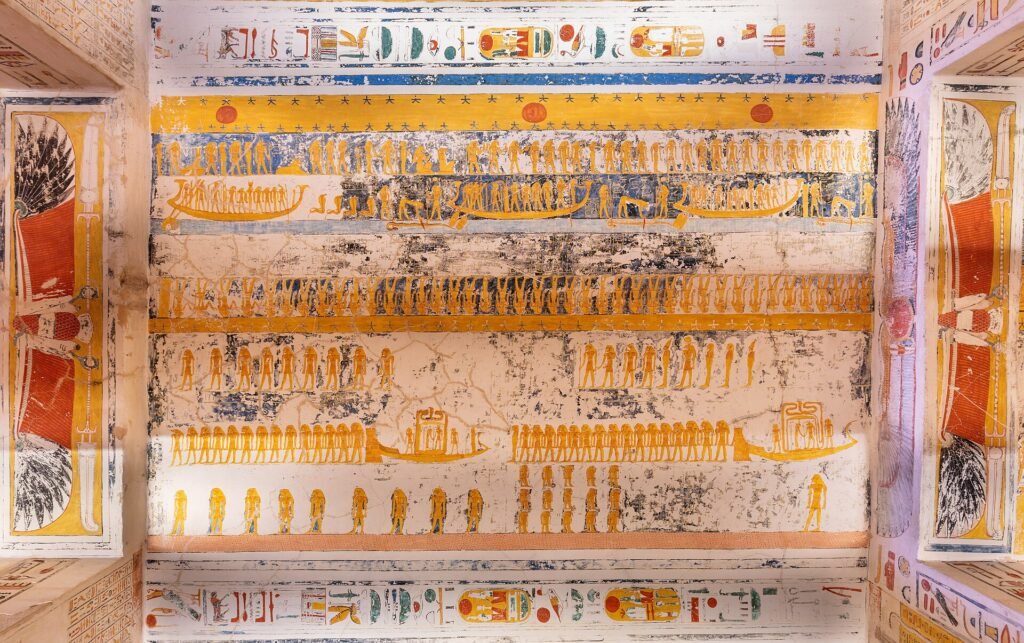 Techo de tumba faraonica en el Valle de los Reyes en Luxor Egipto con filas de figuras doradas y barcas solares pintadas sobre fondo crema y jeroglificos vivos