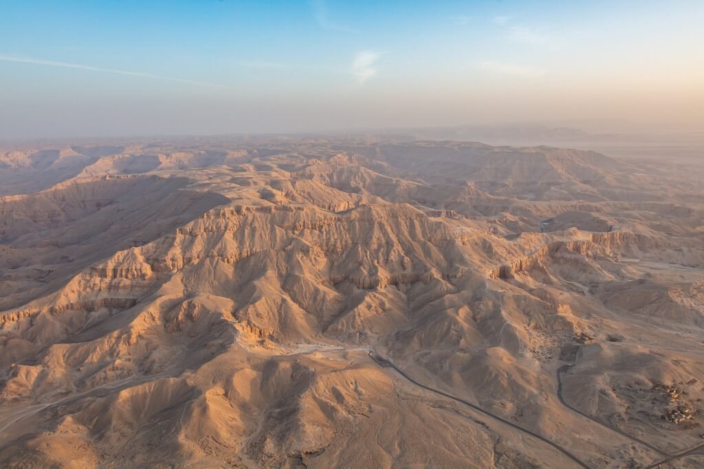 Vista aerea panoramica de las aridas colinas de la necropolis tebana en Luxor al amanecer, con tonos dorados calidos y cielo azul claro, paisaje desertico del valle de tumbas