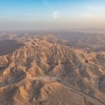 Vista aerea panoramica de las aridas colinas de la necropolis tebana en Luxor al amanecer, con tonos dorados calidos y cielo azul claro, paisaje desertico del valle de tumbas