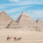 Camello y carruaje cruzando la arena ante las tres piramides de Giza bajo cielo azul
