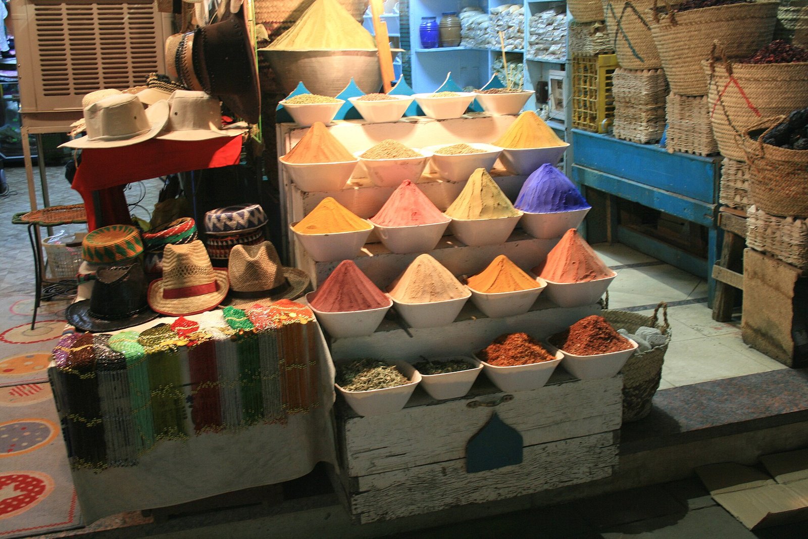 Conos de especias de colores vivos amarillo, naranja y purpura en cuencos blancos junto a sombreros y cestas de mimbre en un mercado tradicional de Egipto