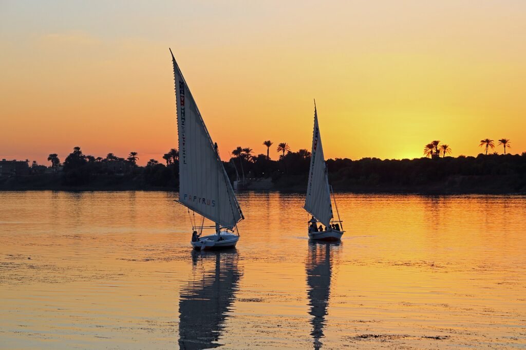 Dos faluccas tradicionales navegan al atardecer sobre el Nilo en Egipto, con el cielo naranja y palmeras reflejadas en el agua, ambiente intimo de grupo reducido