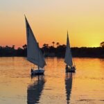 Dos faluccas tradicionales navegan al atardecer sobre el Nilo en Egipto, con el cielo naranja y palmeras reflejadas en el agua, ambiente intimo de grupo reducido