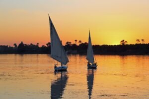 Dos faluccas tradicionales navegan al atardecer sobre el Nilo en Egipto, con el cielo naranja y palmeras reflejadas en el agua, ambiente intimo de grupo reducido
