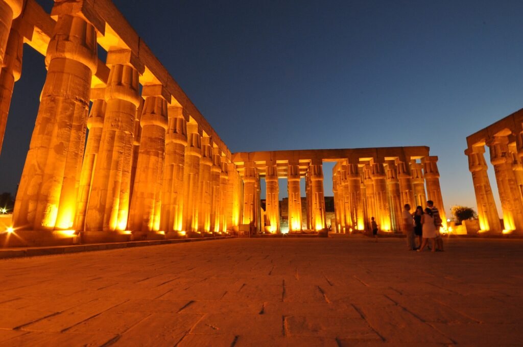 Templo de Luxor iluminado de noche con columnas monumentales en tonos dorados y cielo azul oscuro, escena majestuosa de un viaje a Egipto
