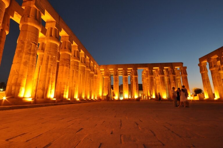 Templo de Luxor iluminado de noche con columnas monumentales en tonos dorados y cielo azul oscuro, escena majestuosa de un viaje a Egipto