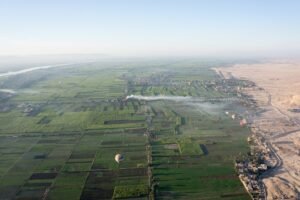 Vista aerea premium del valle del Nilo en Egipto con campos verdes geometricos, el rio brillando y el desierto al borde, con globo aerostatico al amanecer