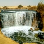 Las cascadas de Wadi El Rayan en El Fayum cayendo sobre un ancho borde hacia una poza verde turquesa, enmarcadas por bancos de arena dorada del desierto egipcio