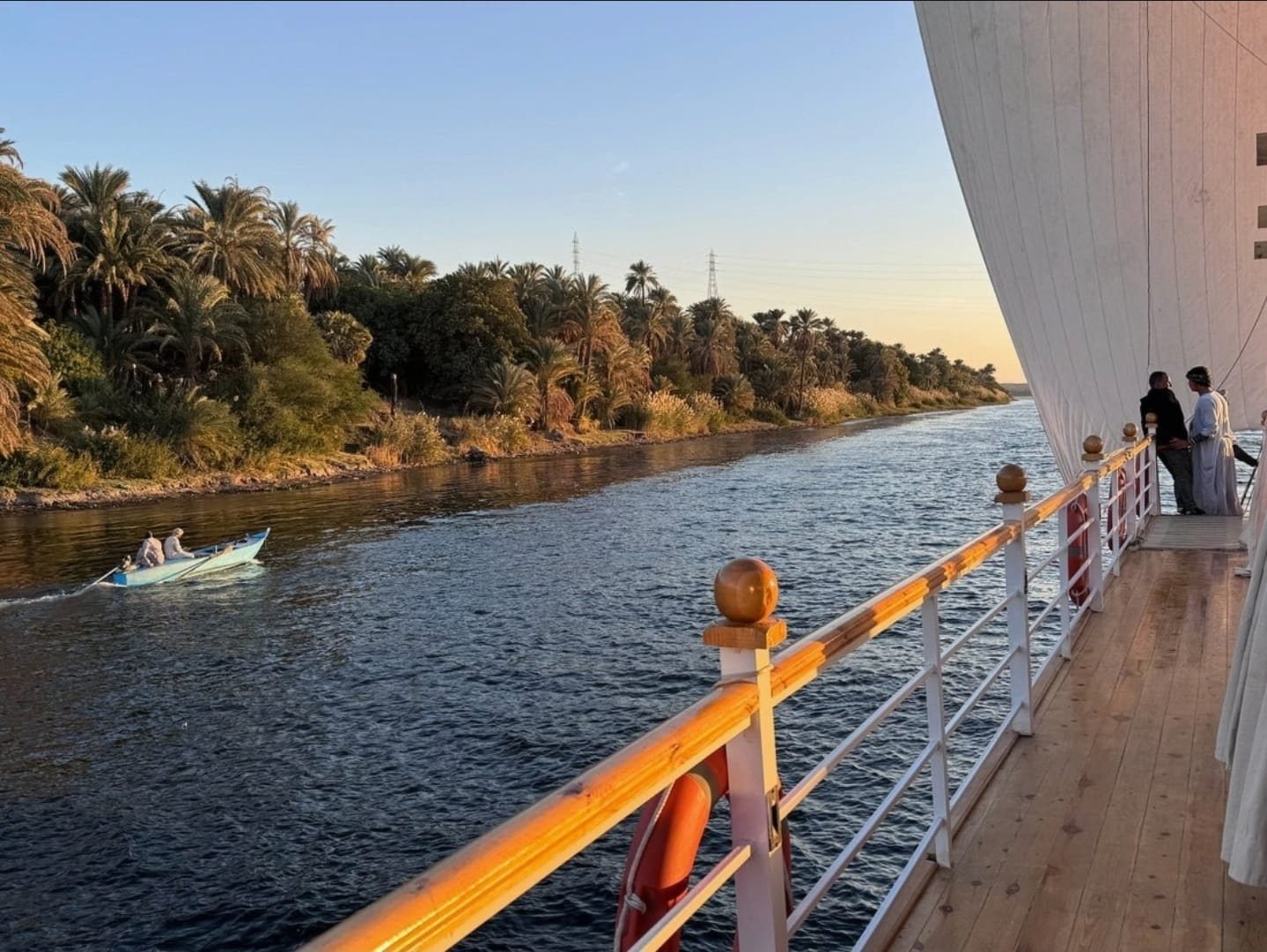 Pasarela lateral de la Akhenaton al atardecer, junto a la vela y una orilla con palmeras