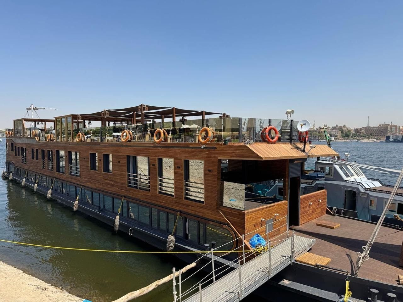 The wooden hull of a dahabiya moored at the riverbank, its upper deck under shade sails.