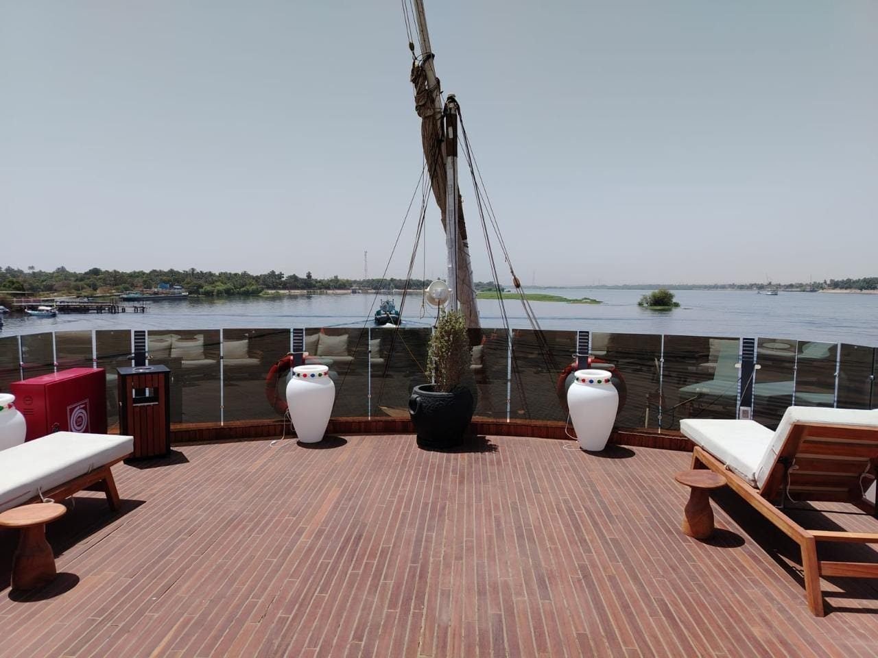 Upper deck of the Iris with sun loungers, white urns and the mast, facing the green bank of the Nile