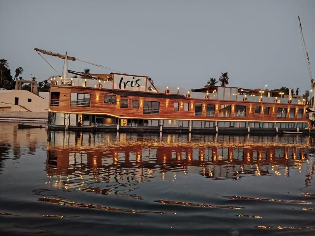 The Iris dahabiya lit up at dusk, moored on the Nile with its lights reflected in the water