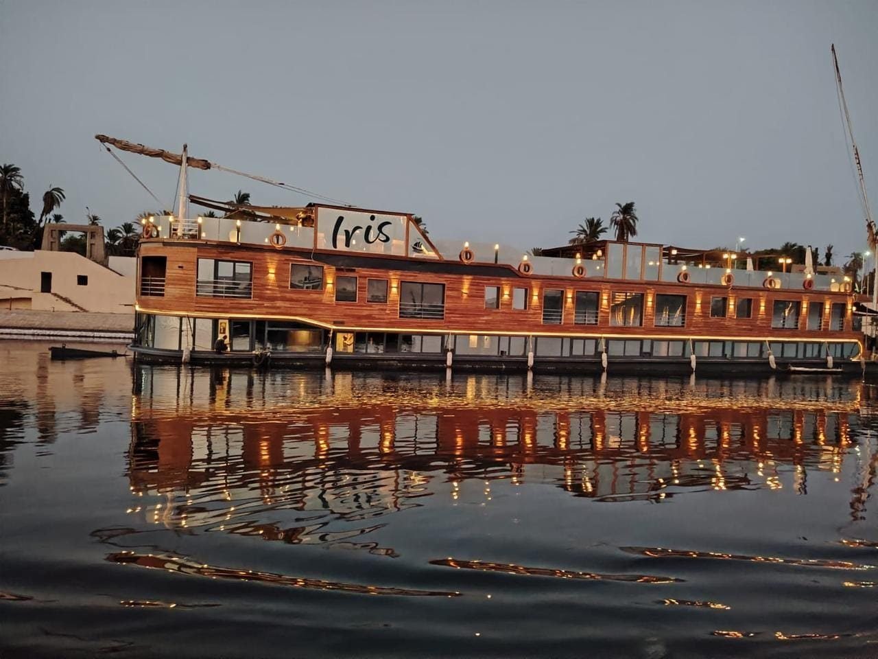 A dahabiya lit up at dusk, moored on the Nile with its lights reflected in the water.
