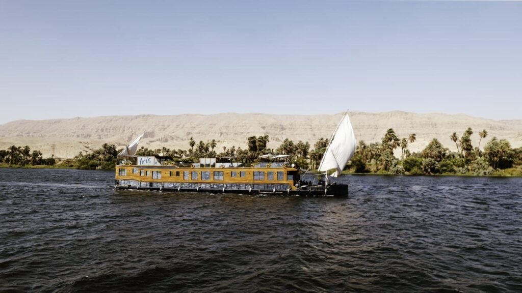 The Iris sailing down the Nile with its white sail up, between a palm grove and desert hills