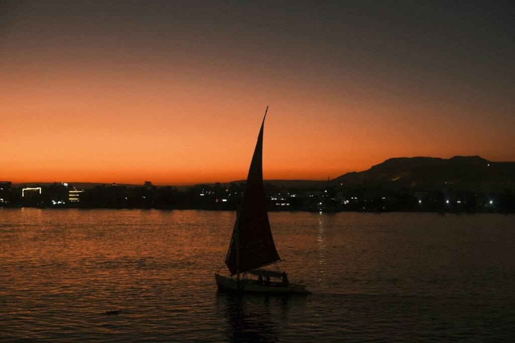La silueta de una faluca con la vela alzada sobre el Nilo al caer el sol, con las colinas de la orilla oeste al fondo