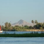 A red-sailed felucca on the Nile at Luxor, with palms and the Theban mountain behind