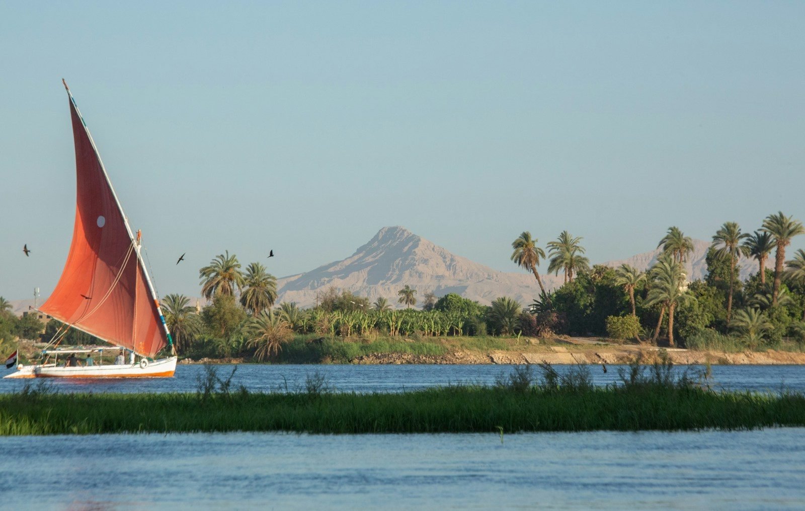 A red-sailed felucca on the Nile at Luxor, with palms and the Theban mountain behind