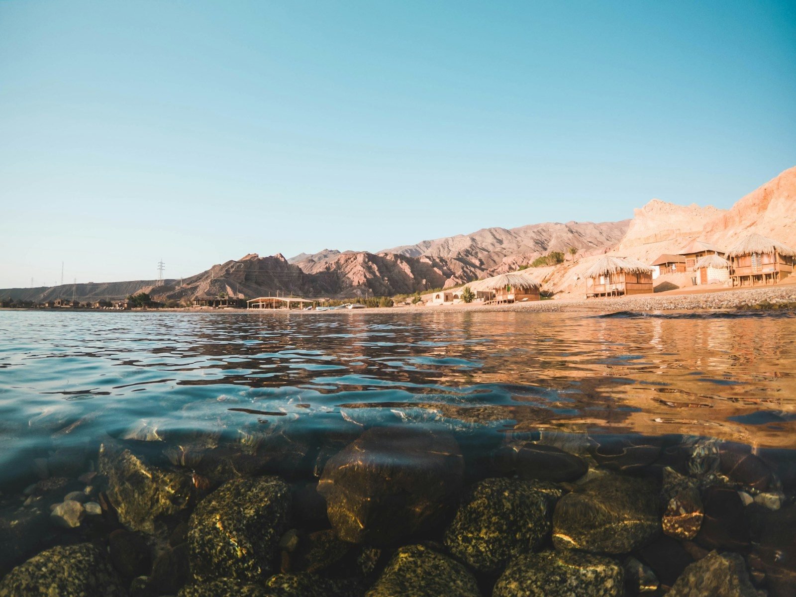 The shore at Nuweiba on the Sinai coast, with clear water over rocks and beach huts below the mountains.