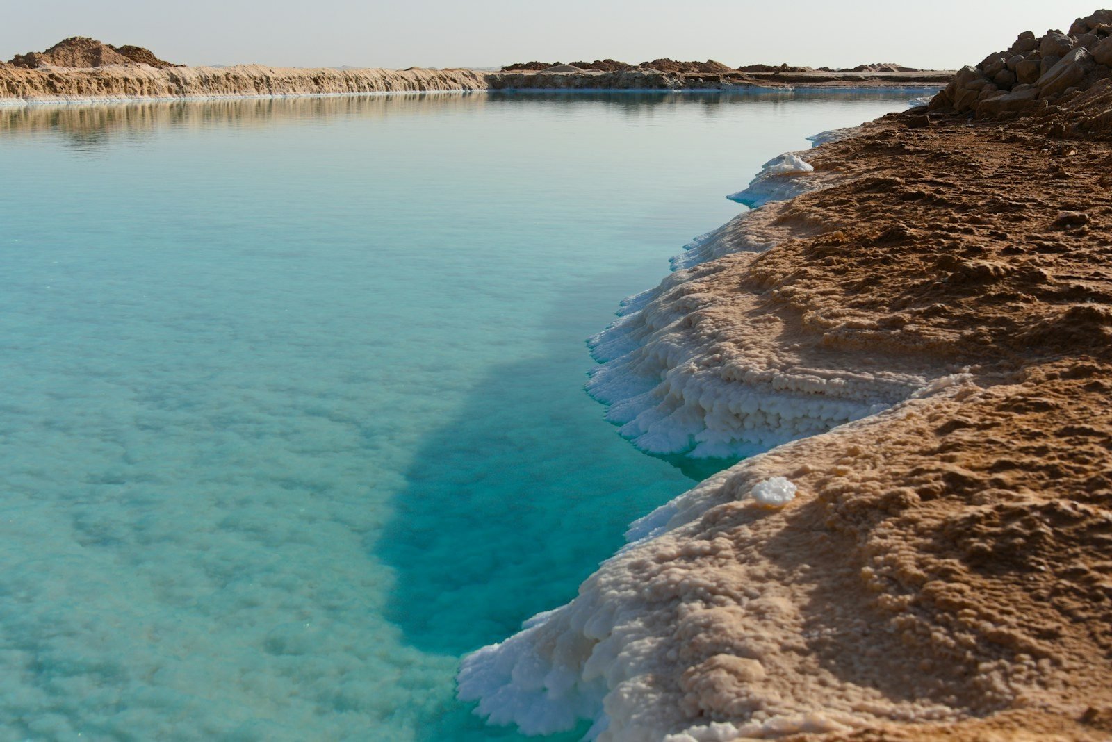 Calm turquoise water beside a brown clay bank edged with a white salt crust.