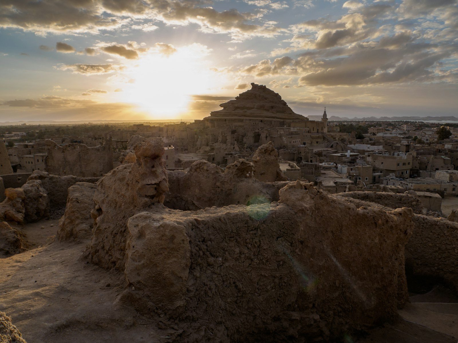 Atardecer sobre las ruinas de adobe de una antigua ciudad de oasis, con una colina de roca escalonada y un minarete en el horizonte y el sol entre las nubes.