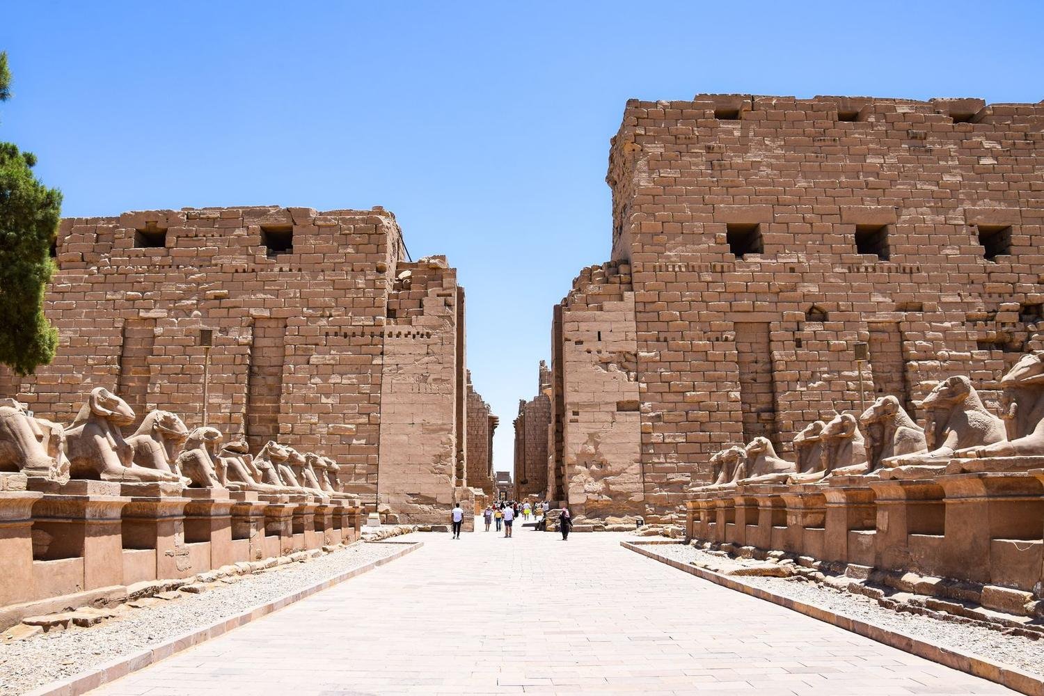 Avenida empedrada flanqueada por esfinges con cabeza de carnero que conduce a los enormes pilonos de piedra de Karnak bajo un cielo azul