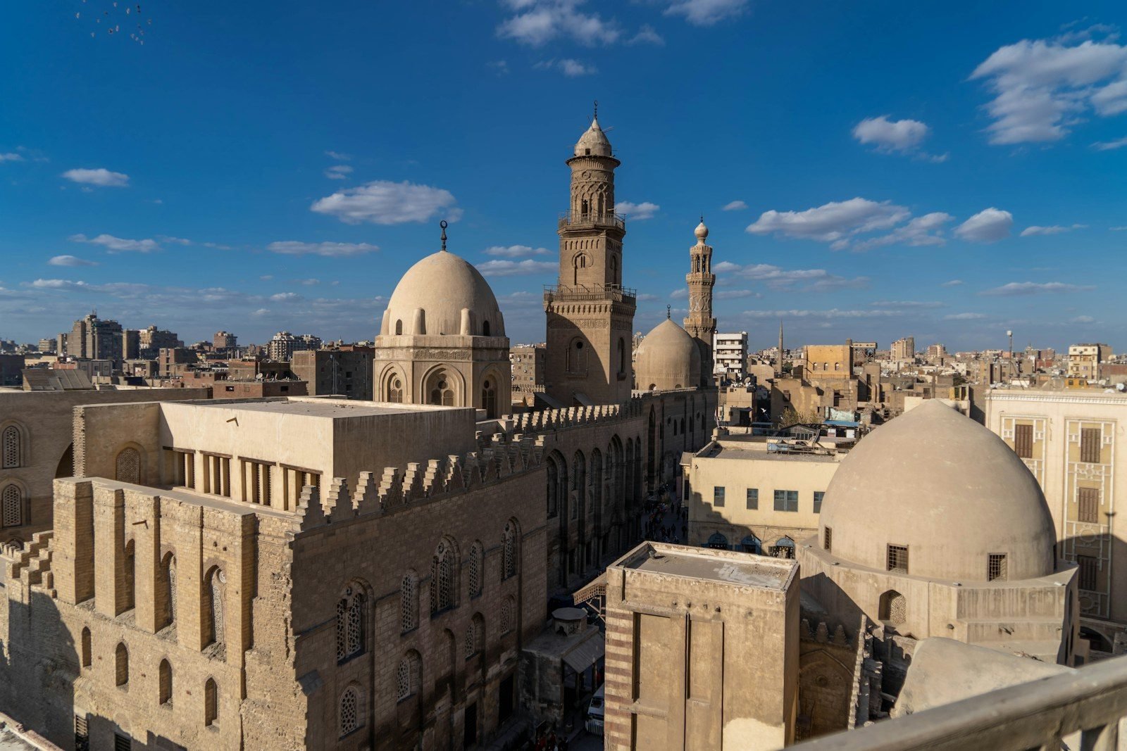 Vista desde una azotea sobre el Cairo islámico, con cúpulas de piedra, dos minaretes labrados y muros almenados sobre una calle estrecha, bajo un cielo azul con nubes dispersas.