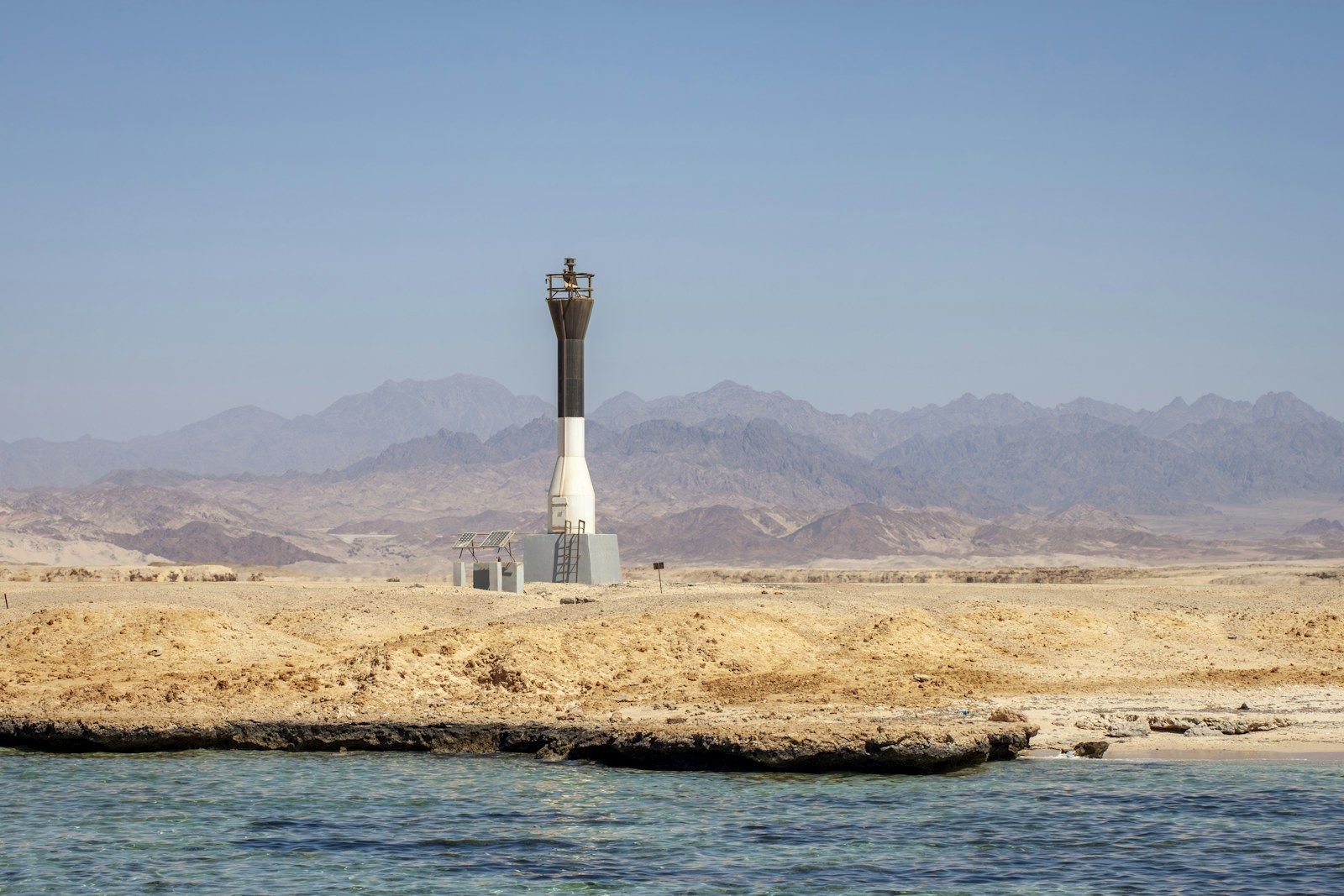 Black and white navigation beacon with solar panels on a rocky desert shore, with arid mountains behind and turquoise water in front.