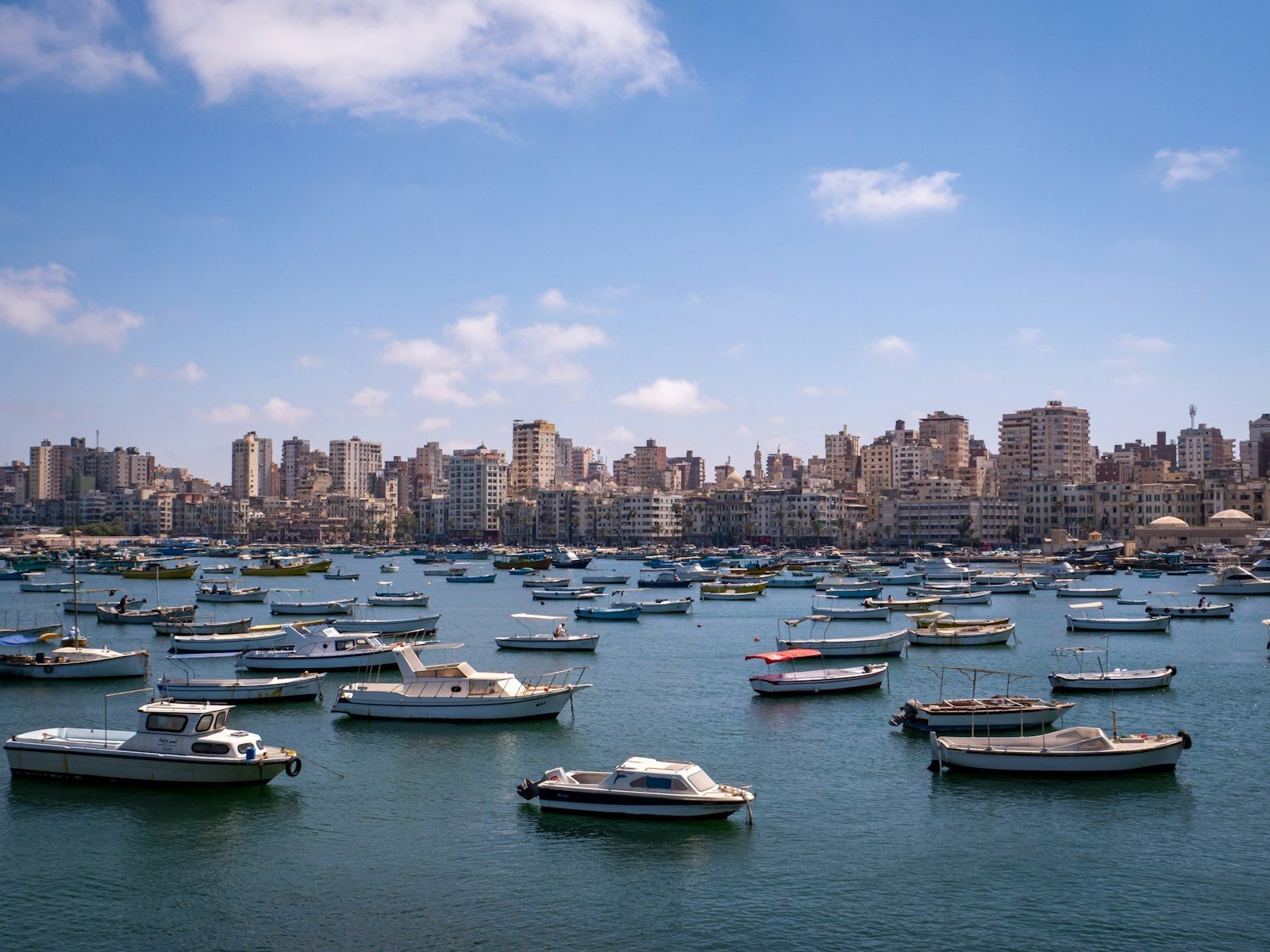 Numerosas barcas blancas y pequeñas lanchas amarradas en aguas tranquilas de un puerto, con una línea de edificios de apartamentos y alguna cúpula al fondo bajo un cielo azul con nubes.