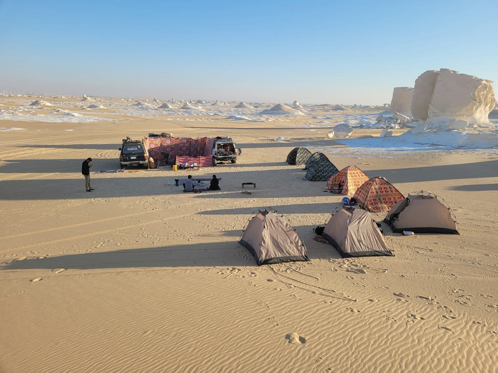 Campamento en el desierto con tiendas de campaña y dos todoterrenos junto a un cortavientos de tela, con bloques de roca blanca al fondo bajo un cielo despejado.