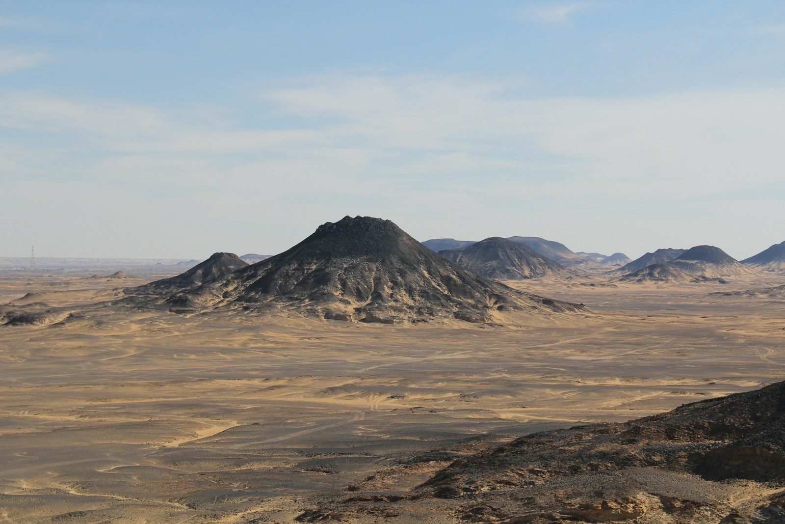 A dark conical hill and a chain of black hills rising from a pale sand plain under a lightly hazy sky.
