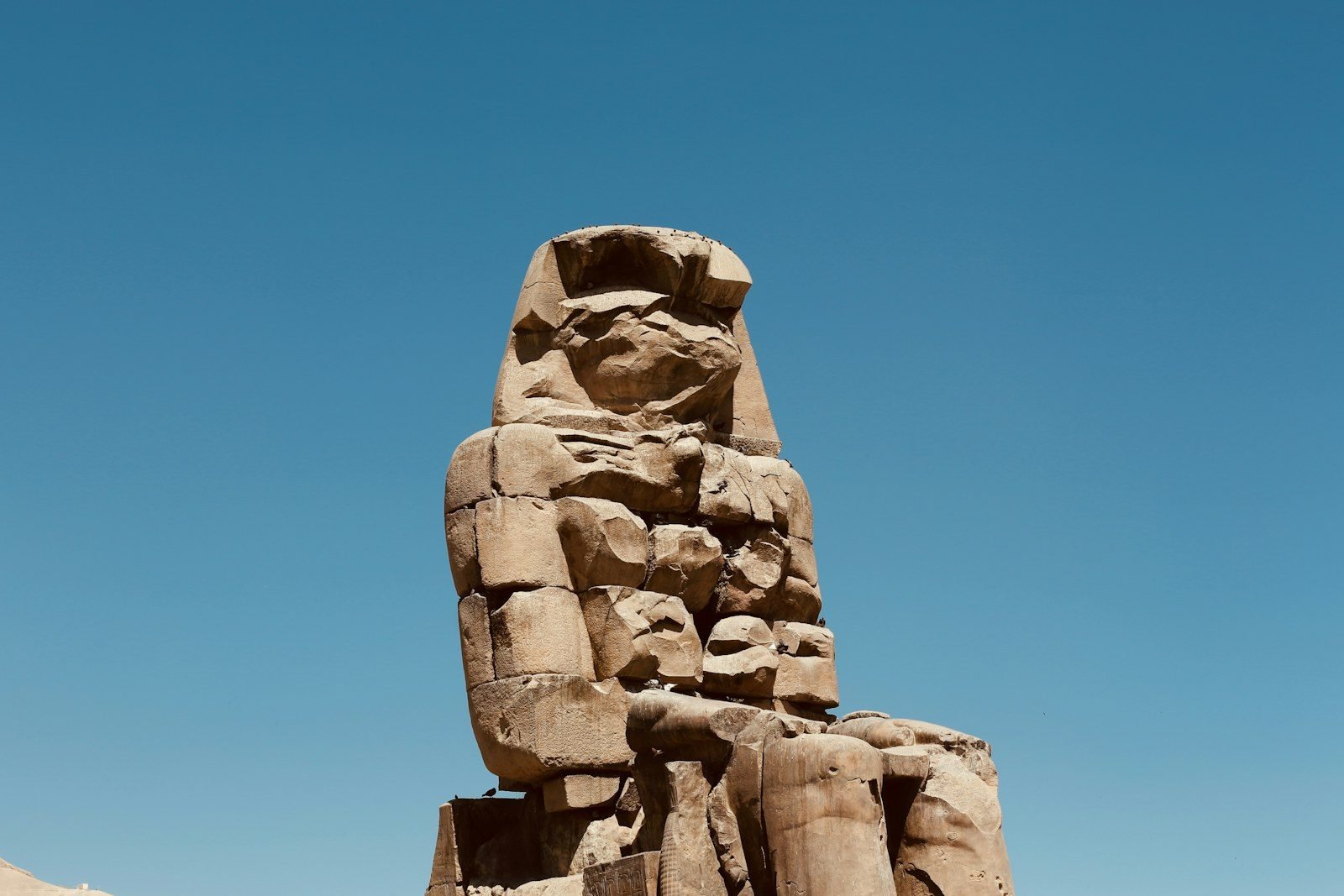 Primer plano de uno de los colosos de Memnón, una estatua sentada de piedra agrietada, recortada contra un cielo azul sin nubes.