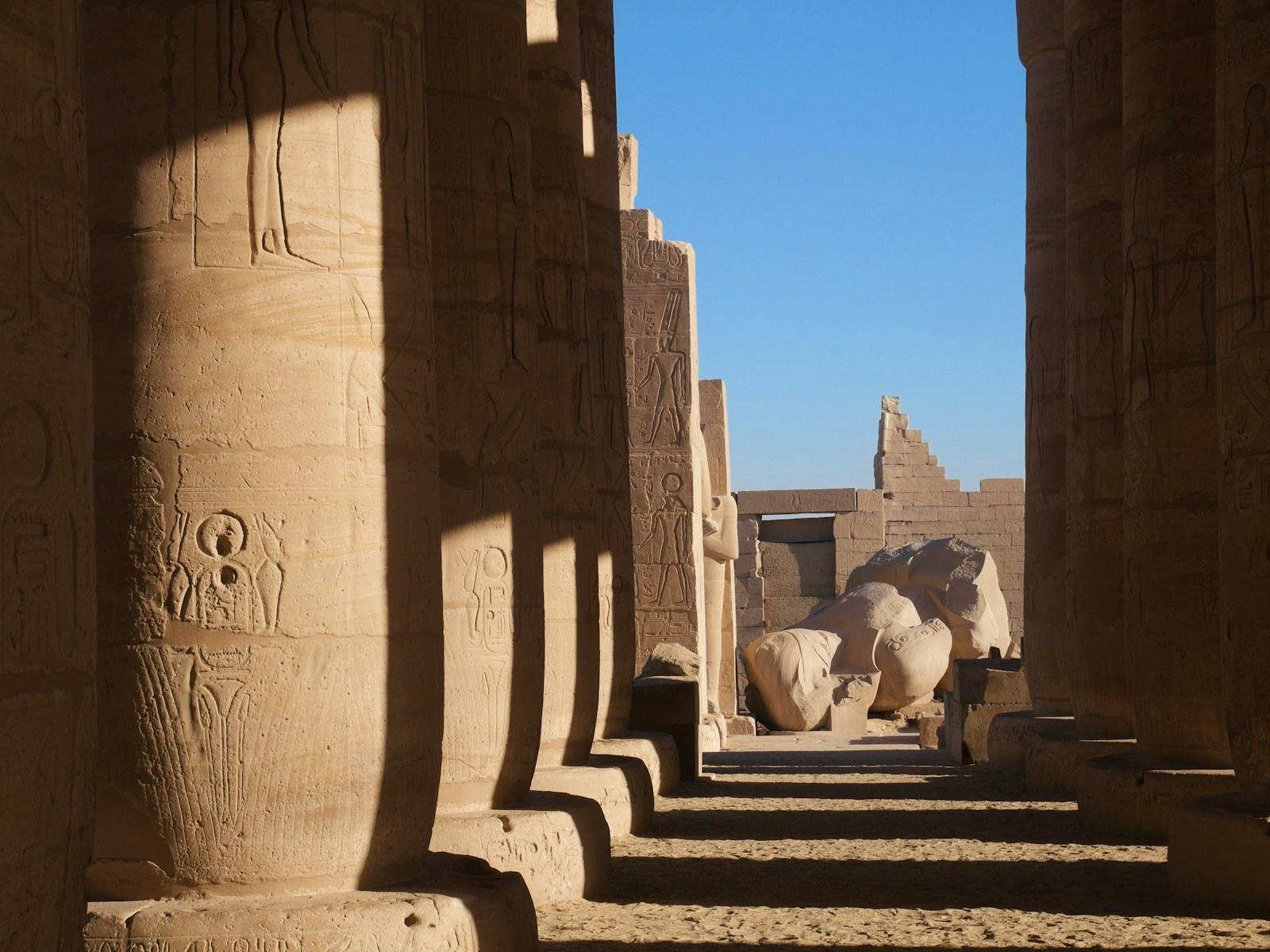 Columnas de arenisca con relieves enmarcan una estatua colosal caída y un muro escalonado en la sala de un templo antiguo bajo sol intenso