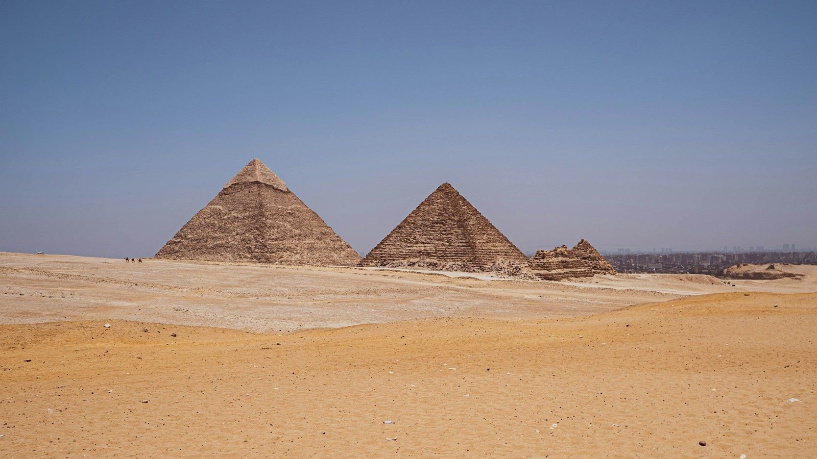 Dos pirámides de piedra de distinto tamaño y una pirámide menor se elevan sobre la arena dorada del desierto, con una ciudad lejana entre la bruma