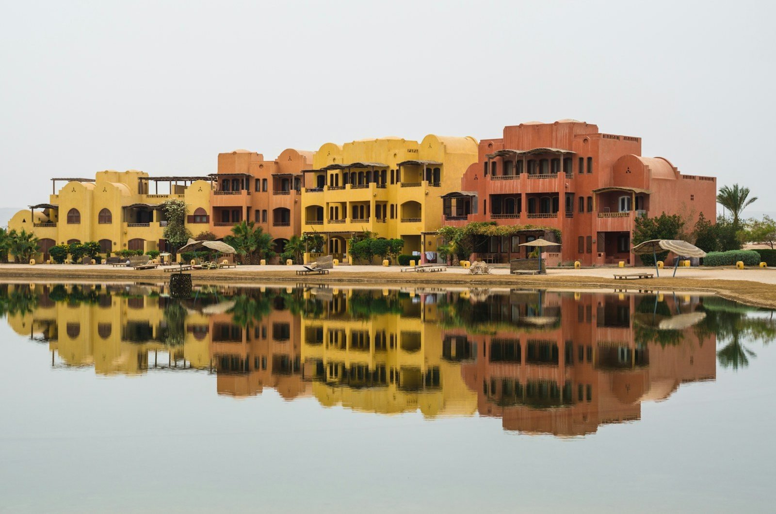 Edificios de tres plantas en tonos ocre, amarillo y terracota, con balcones y arcos, reflejados en el agua quieta de una laguna, con sombrillas de paja en la orilla.