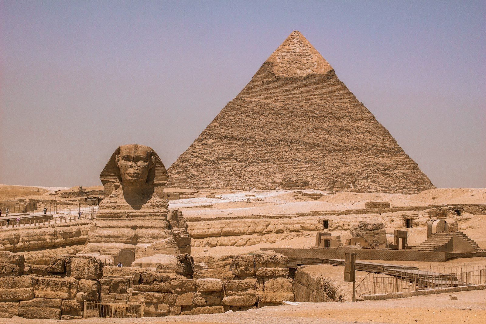La Gran Esfinge de Giza vista de frente con la pirámide de Kefrén detrás, sillares en ruinas en primer plano y las vallas del recorrido, bajo un cielo violáceo con bruma.