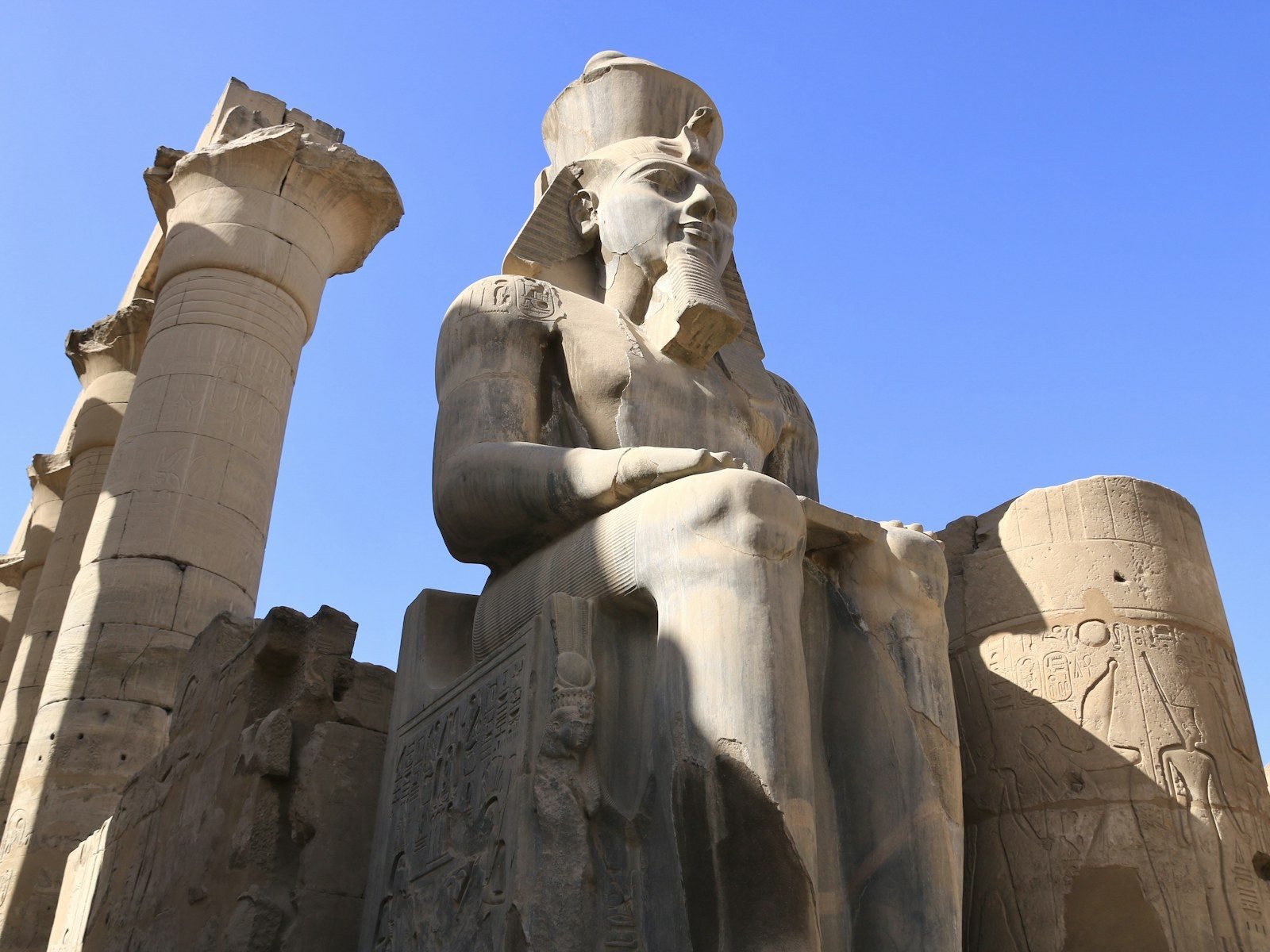 Estatua colosal de un faraón sentado, vista desde abajo contra un cielo azul, junto a una columna de capitel de papiro abierto.