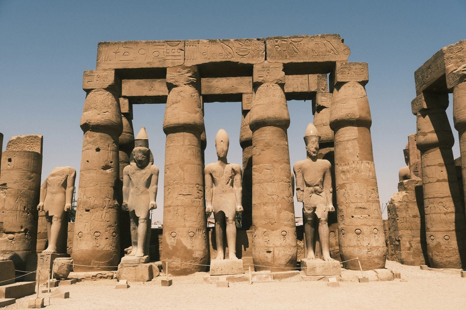 Estatuas de pie de un faraón entre gruesas columnas papiriformes de capitel cerrado en el patio de un templo, con cuerdas que protegen el suelo.