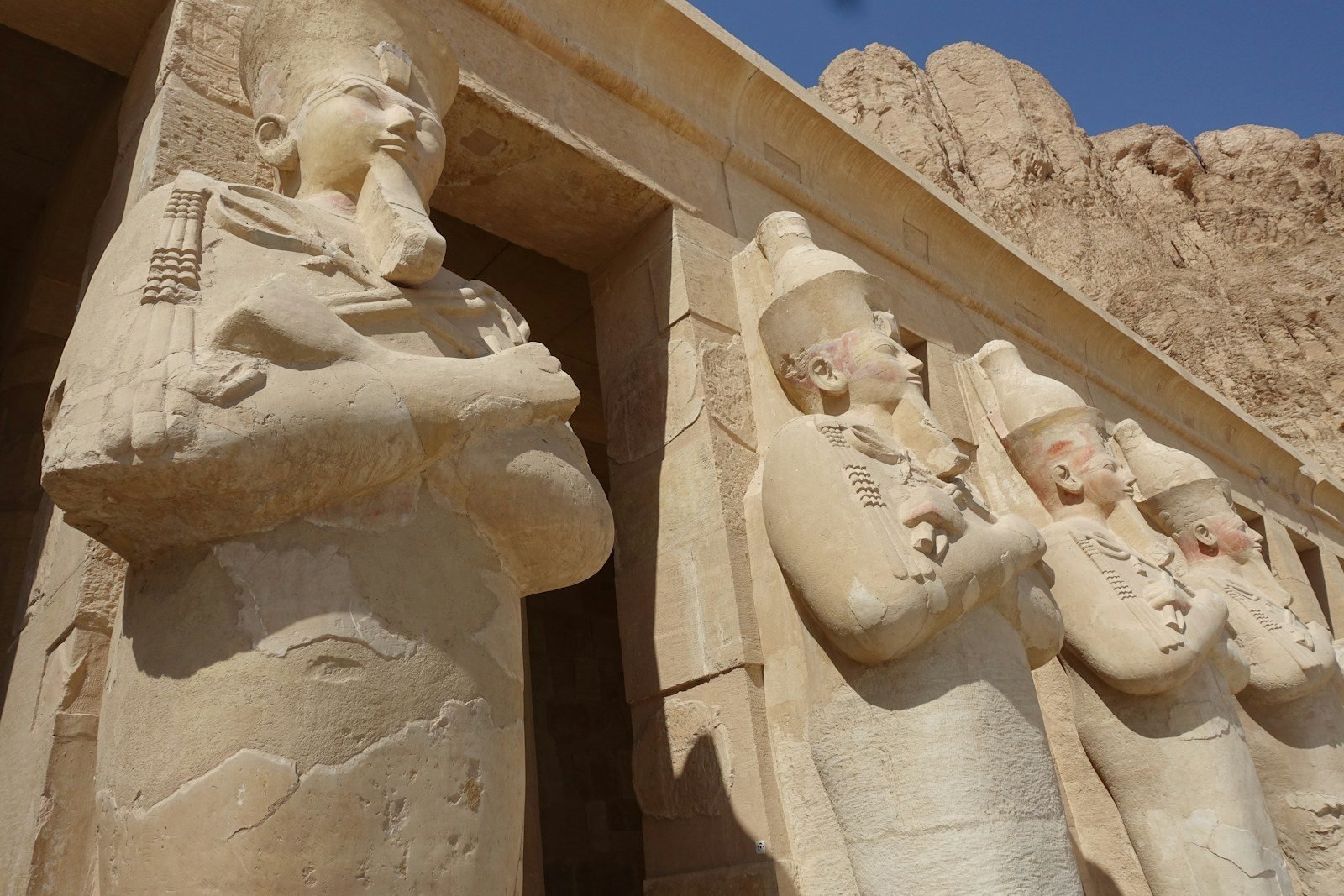 A row of Osiride statues wearing crowns, arms crossed, beside a doorway of the temple of Hatshepsut, with the cliff behind.
