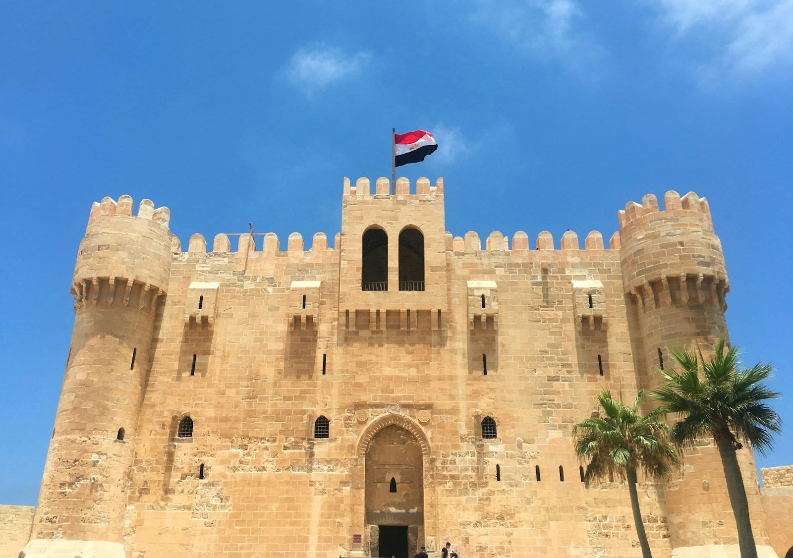 La ciudadela de Qaitbay vista de frente, con sus dos torres redondas en las esquinas, la bandera egipcia y palmeras.