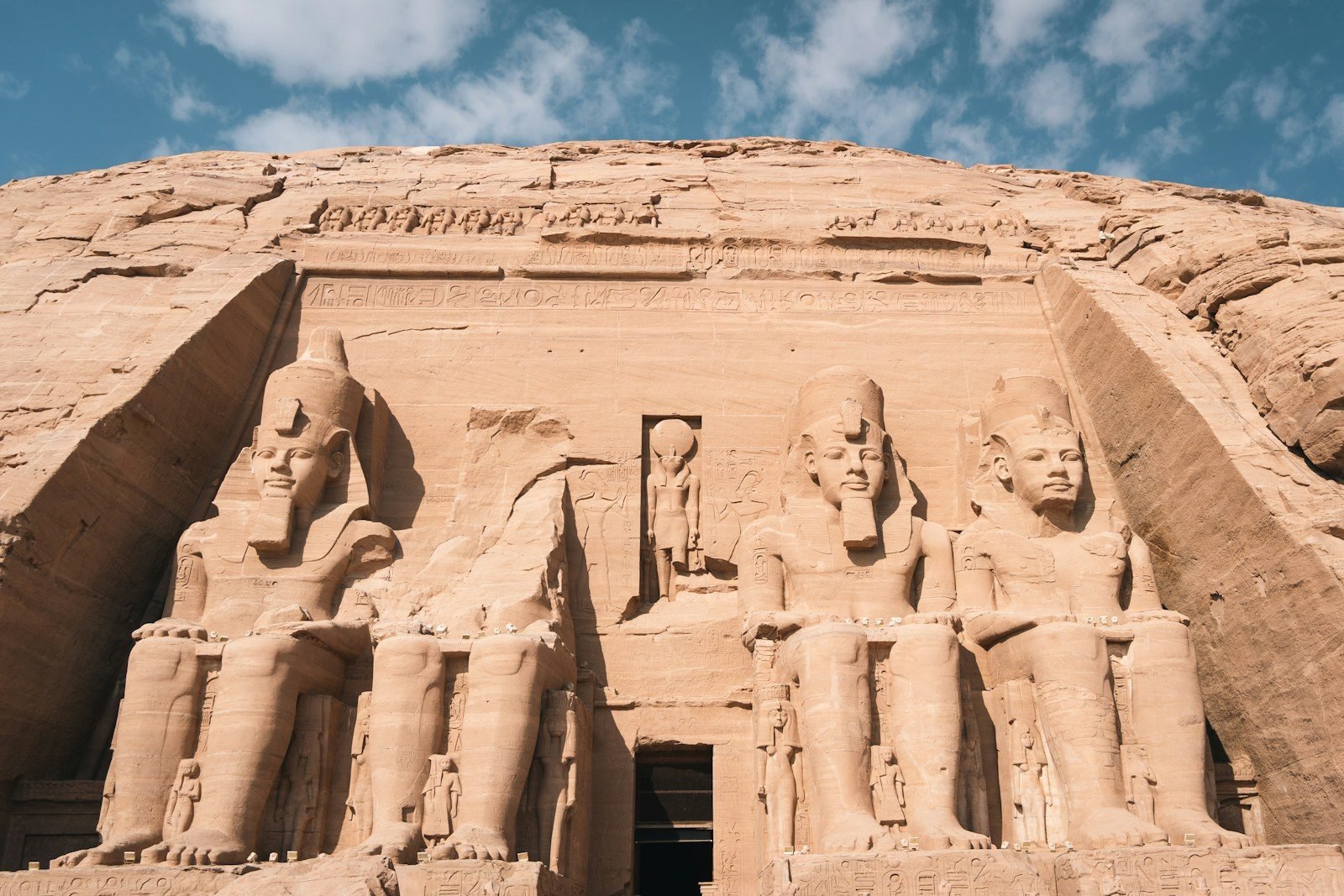 Fachada del gran templo de Abu Simbel vista desde abajo, con los colosos sentados de Ramsés II a ambos lados de la puerta central, bajo un cielo azul con nubes.