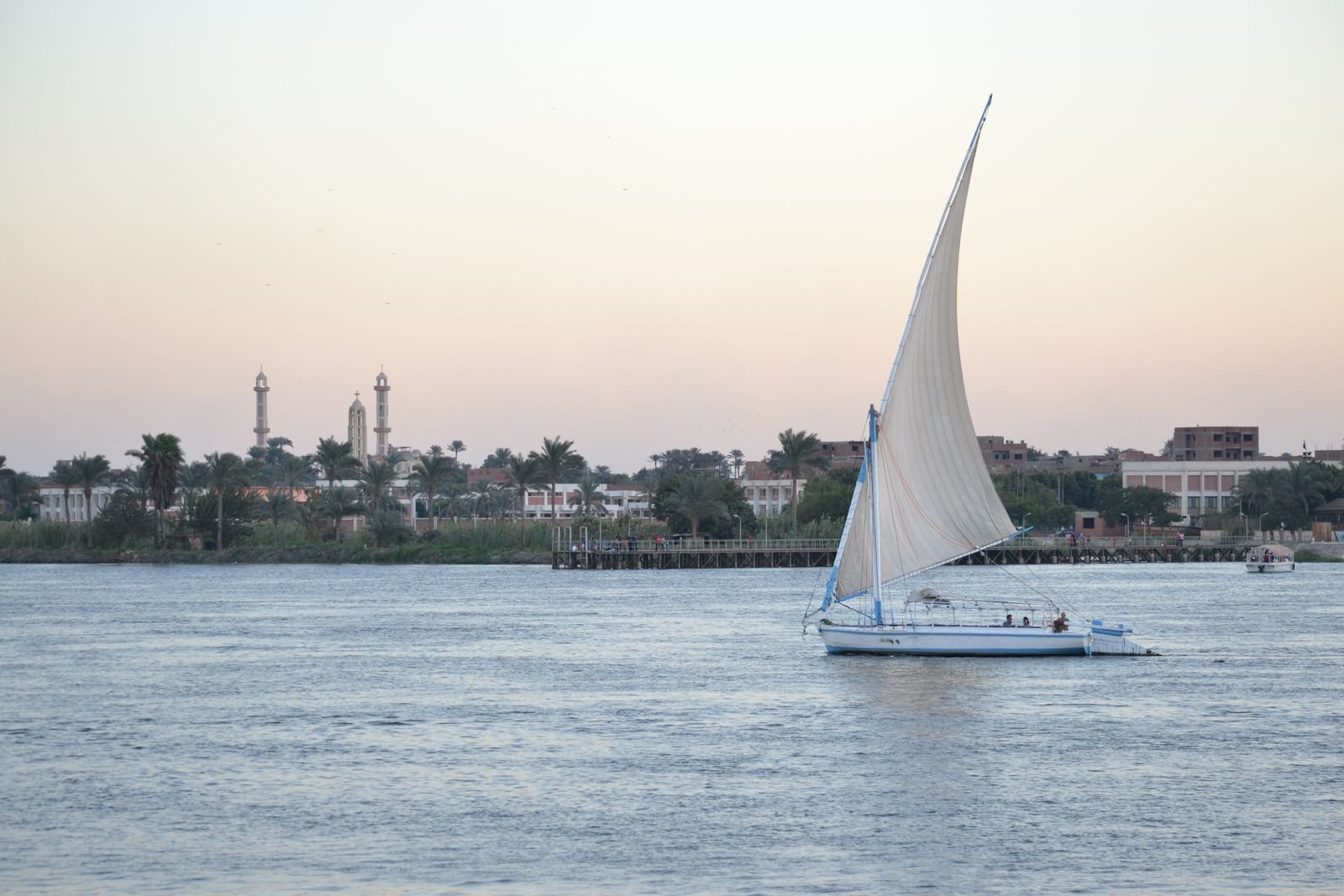 Faluca blanca de vela alta sobre las aguas tranquilas del Nilo al atardecer, con palmeras, edificios y torres esbeltas en la orilla bajo un cielo rosado