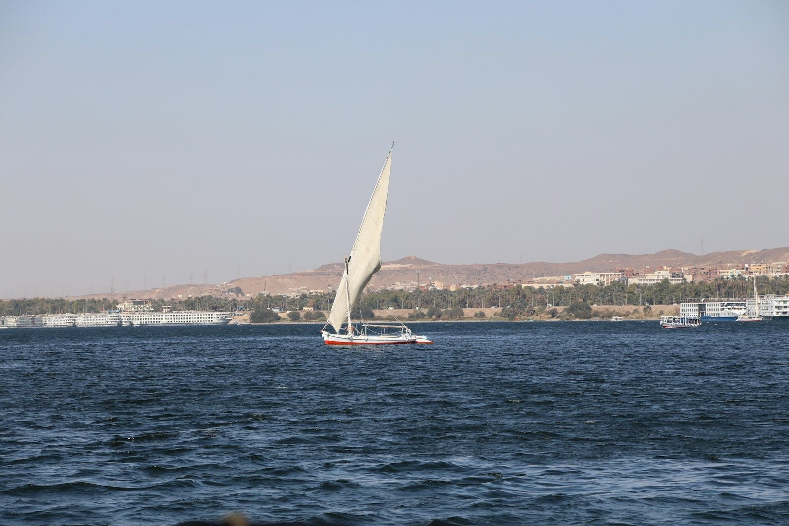 Una faluca de vela triangular clara navega por aguas azul oscuro, con barcos fluviales atracados y colinas áridas en la orilla opuesta