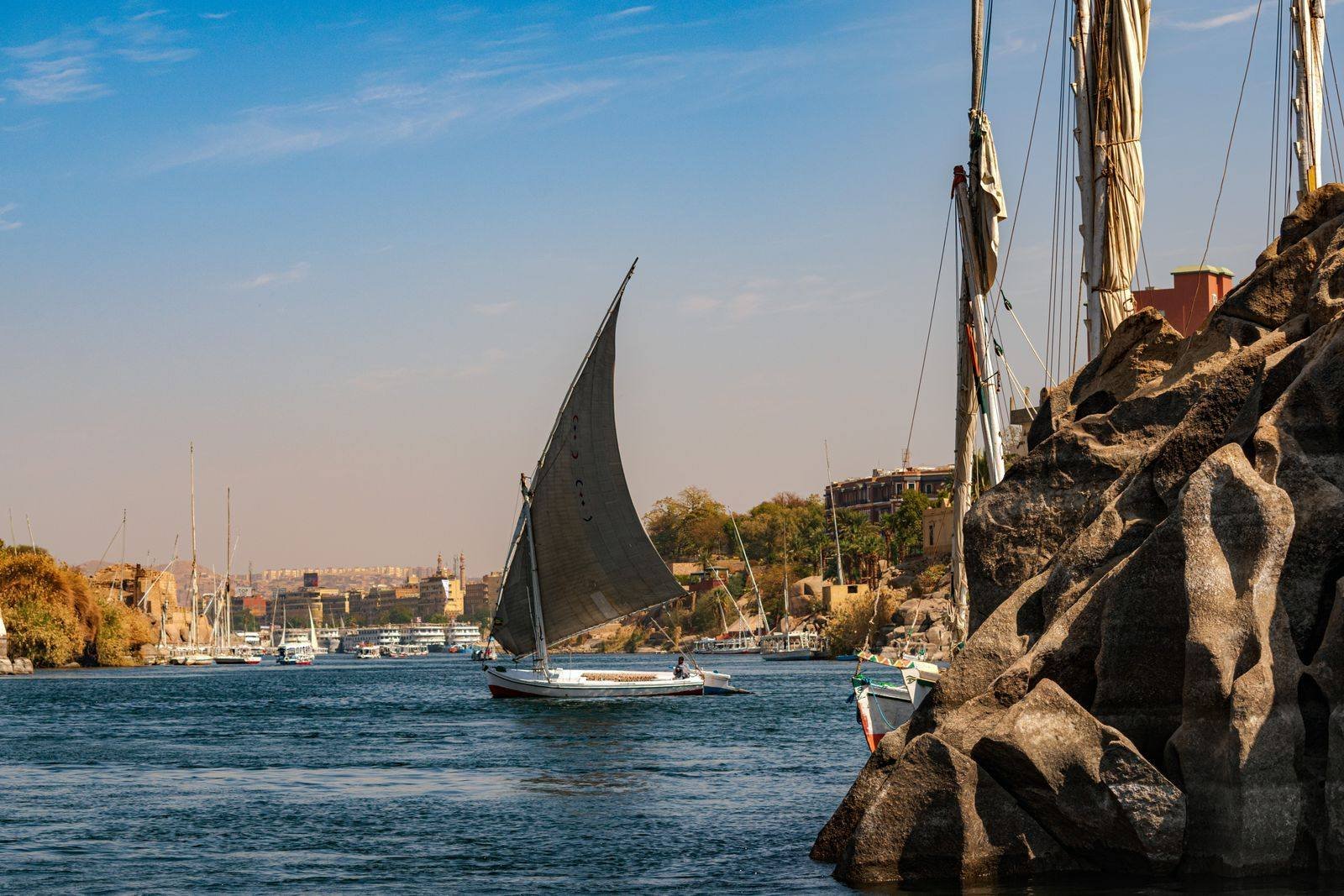 Una faluca con vela triangular gris navega por el Nilo junto a grandes rocas de granito, con barcos amarrados y edificios al fondo en luz dorada
