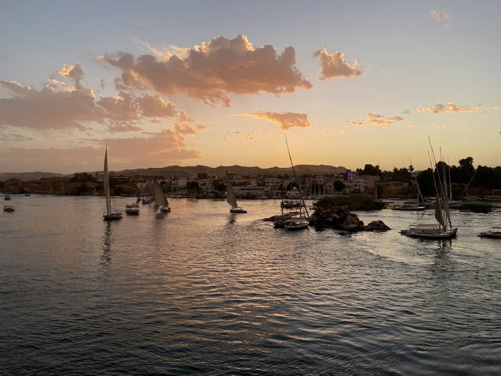 Faluchos navegando y amarrados en un tramo ancho del Nilo al atardecer, con una isla rocosa, un pueblo de casas blancas junto al río y nubes iluminadas por el sol.