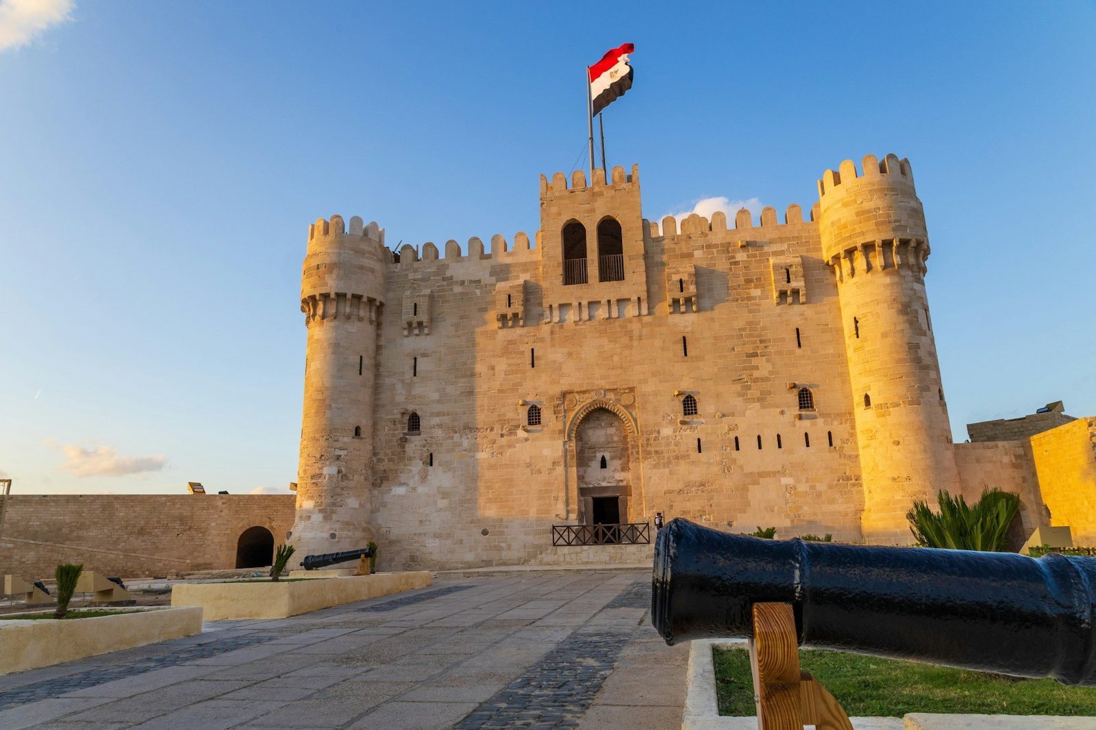 La Fortaleza de Qaitbay iluminada por la luz dorada de la tarde, con la bandera egipcia en lo alto y un cañón negro en primer plano.
