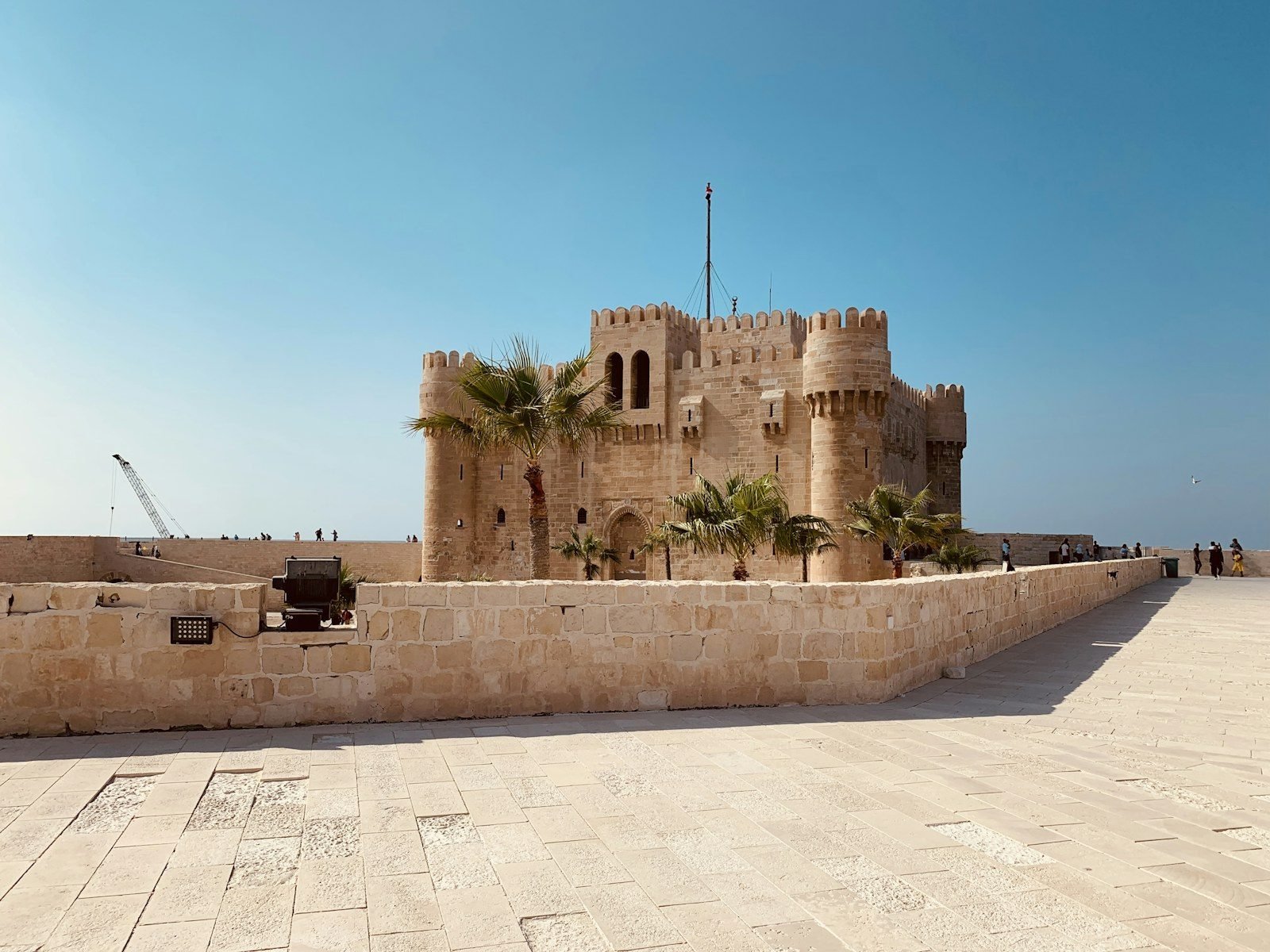 La fortaleza de Qaitbay en Alejandría, con su torre del homenaje almenada y sus torres redondas entre palmeras, vista sobre un muro de piedra caliza bajo un cielo azul limpio.
